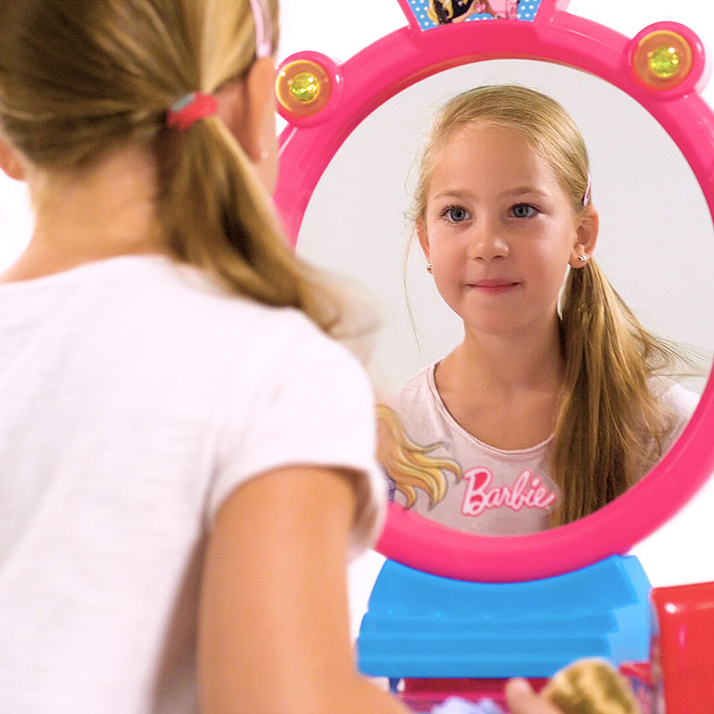 Blick an einem Mädchen mit langen blonden Haaren vorbei auf einem Spiegel, in dem es sich selbst ansieht. Der Spiegel gehört zu einem Schminktisch der in diesem Produktvideo beworben wird.