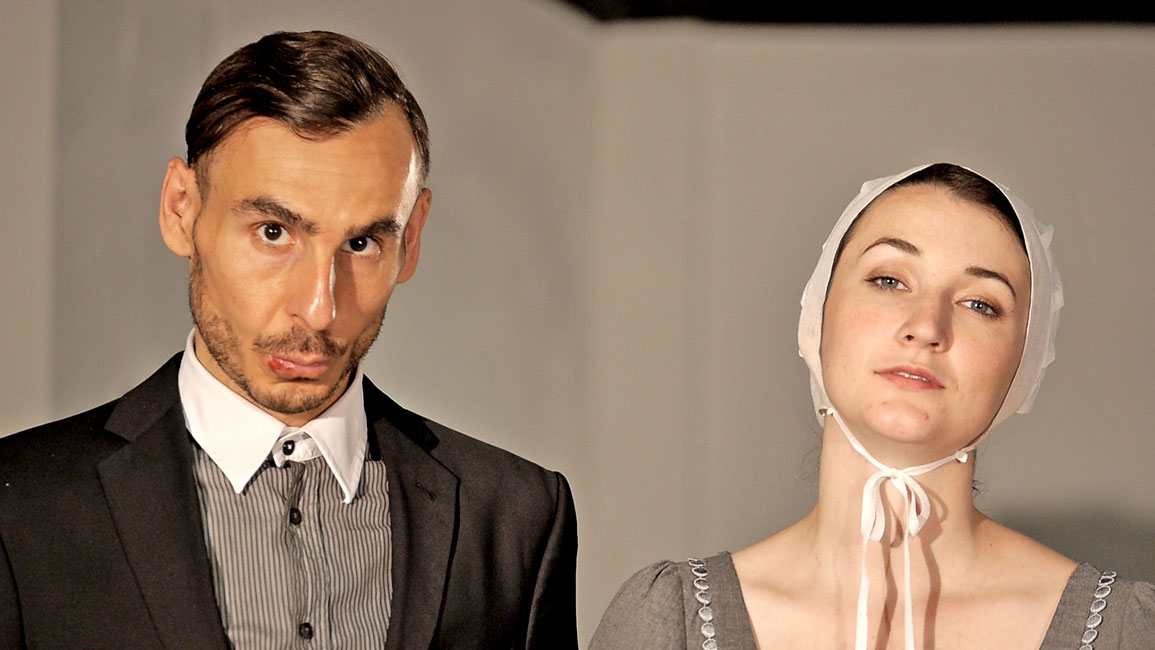 Einstieg in den Theater Trailer: Eine Frau und ein Mann stehen nebeneinander. Beide schauen mit ernstem Blick in die Kamera. Die Kleidung entstammt nicht diesem Jahrhundert.